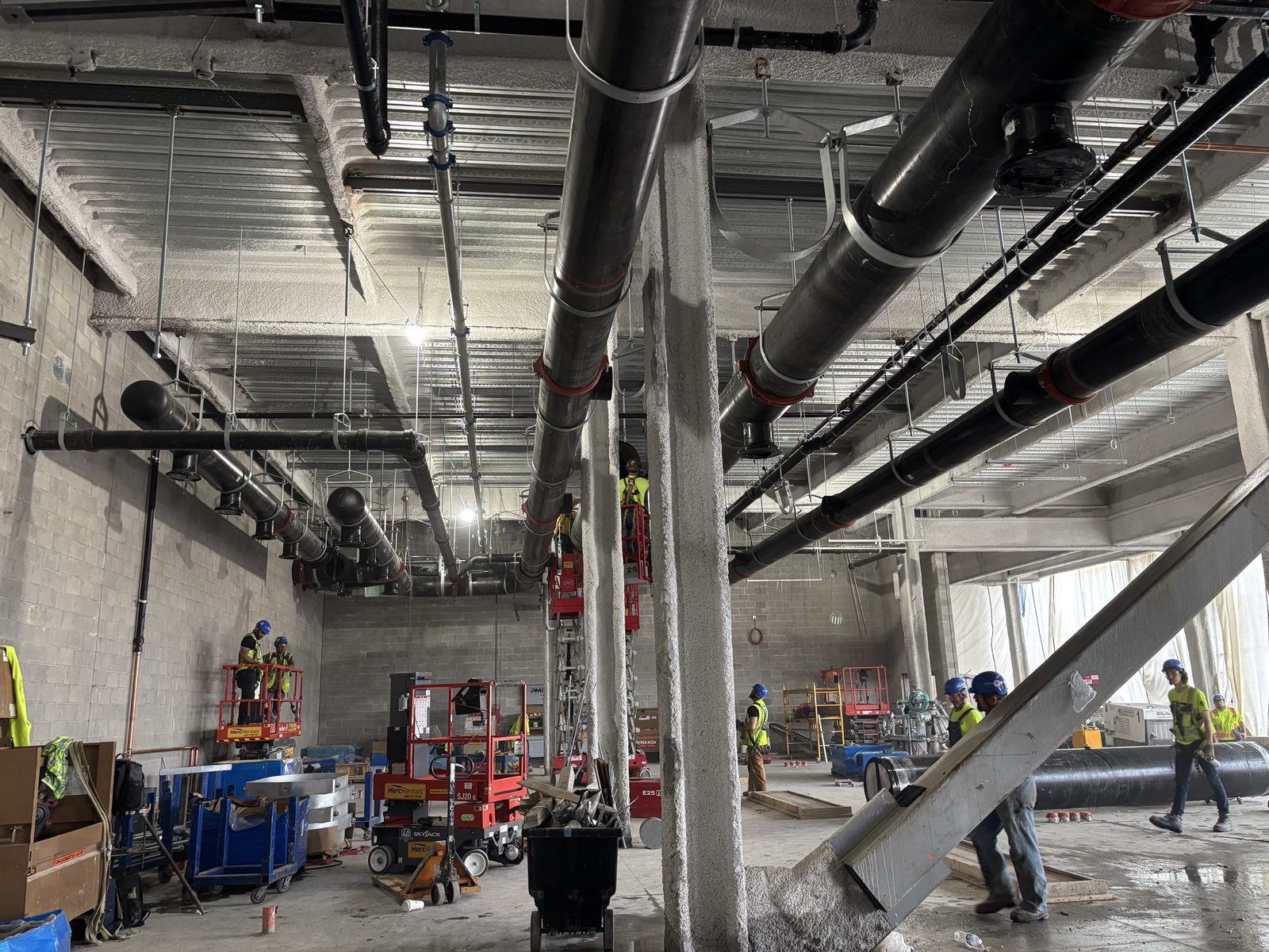 A commercial mechanical room mid-build with overhead pipe runs and equipment staged on the floor